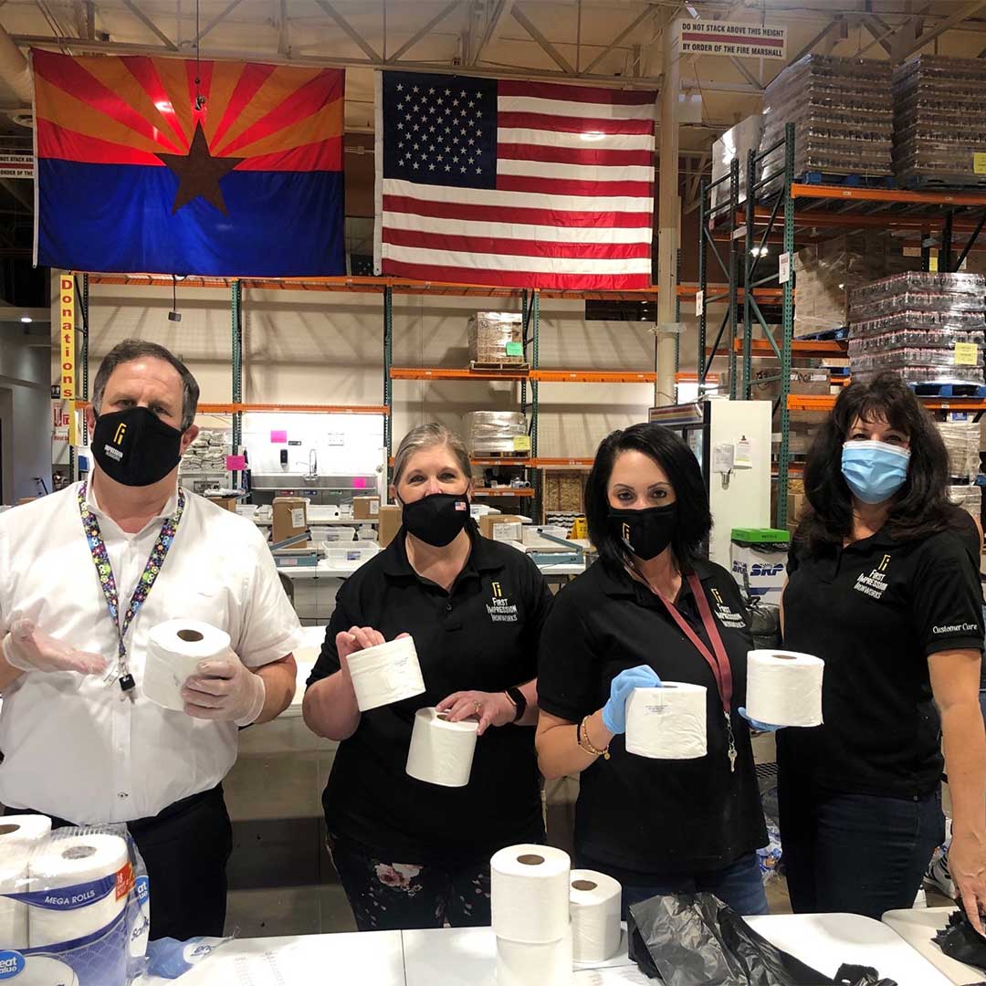 First Impression Ironworks team members holding multiple rolls of toilet paper that are being packed into donation boxes